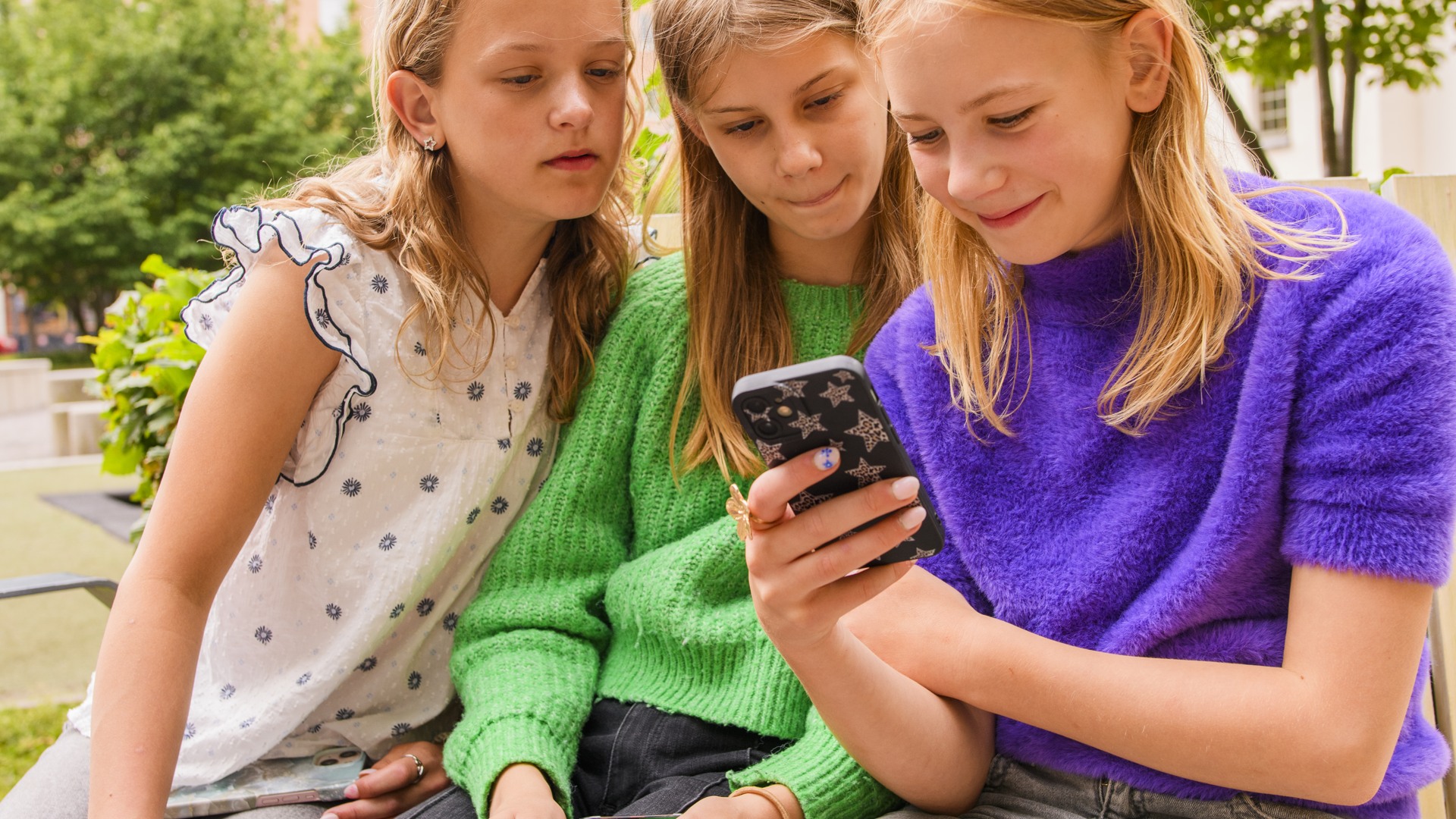 Tre flickor sitter p&aring; en b&auml;nk och tittar p&aring; en mobiltelefon.