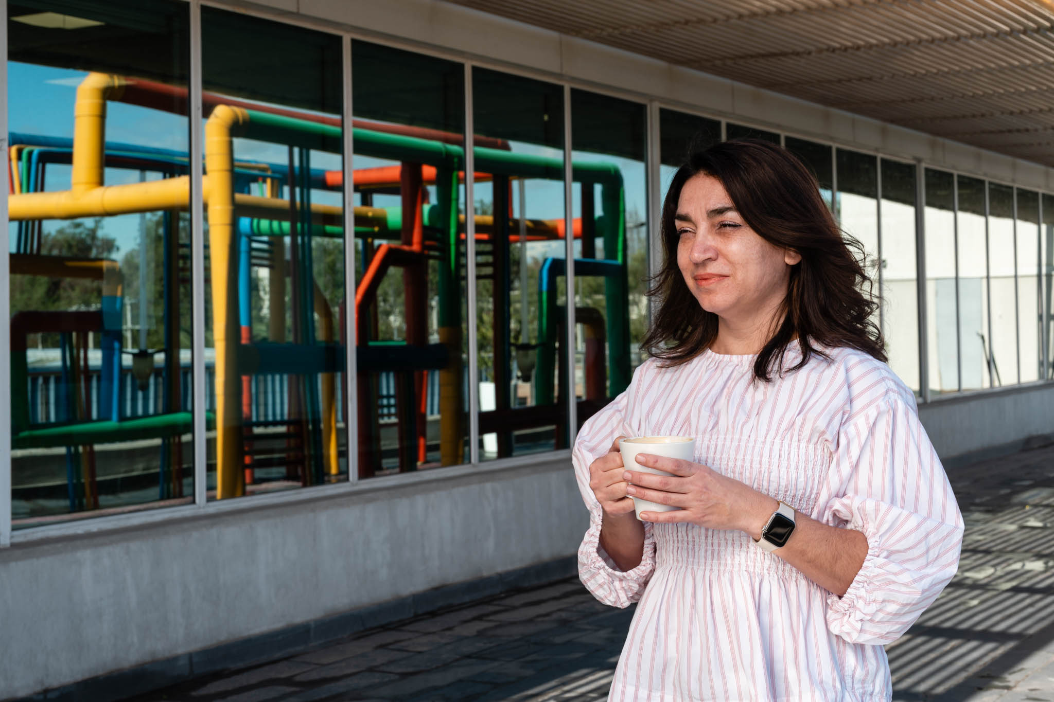 Pia Quiroz, dressed in a pink and white-striped top, walks outside carrying a coffee mug. The colorful pipes from the Google data center are reflected in the windows as she passes.